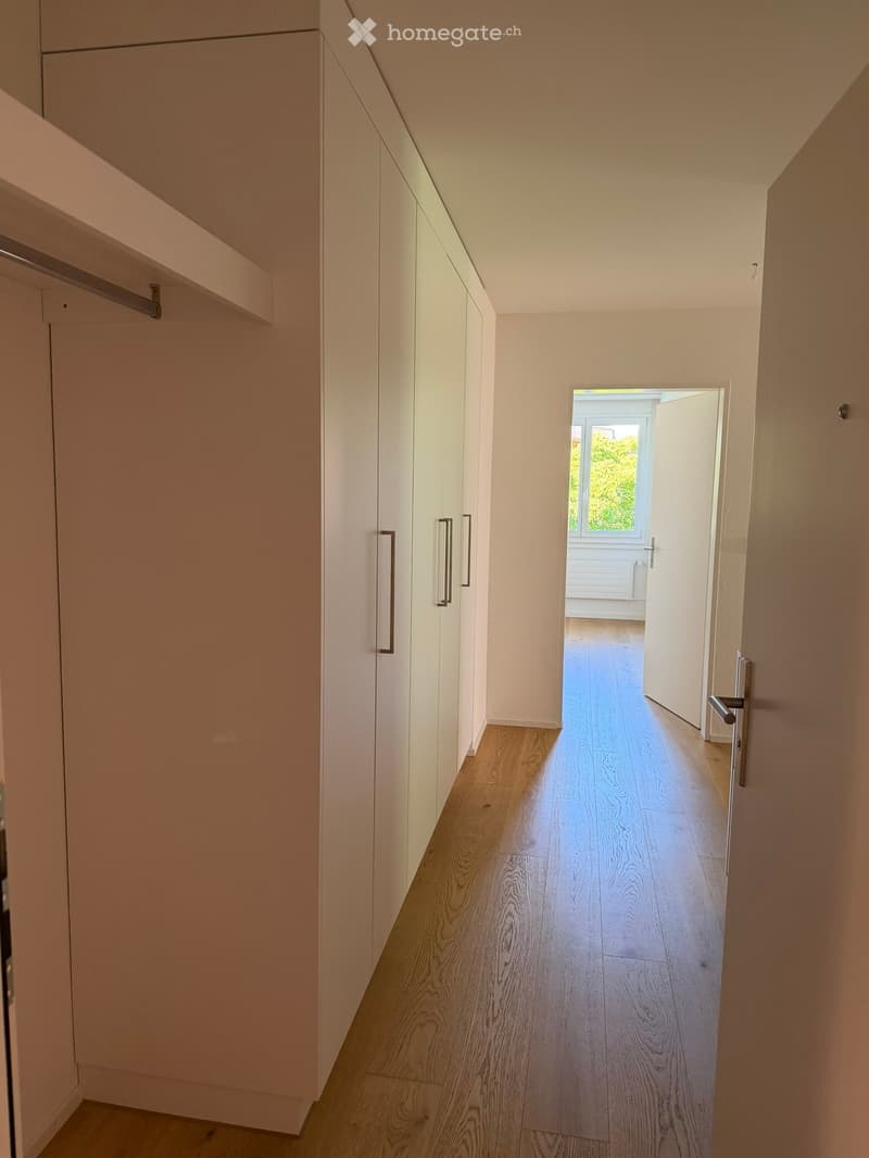 A modern hallway with light wooden flooring, white walls, and built-in white cabinets with sleek handles. An open doorway leads to another room with visible natural light and greenery outside.