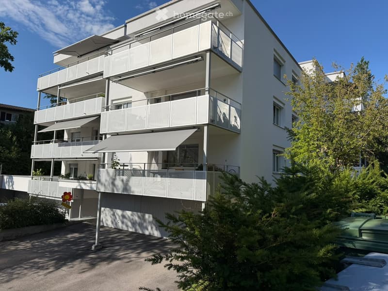 Modern multi-story residential building with white exterior, multiple balconies, and large windows. Features include a covered terrace area and surrounding greenery.