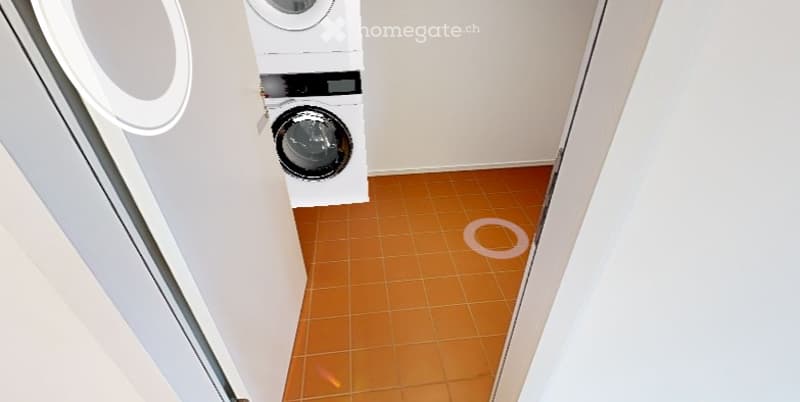 Empty room with orange tiled floor and white walls, featuring a washing machine and dryer unit in the corner.