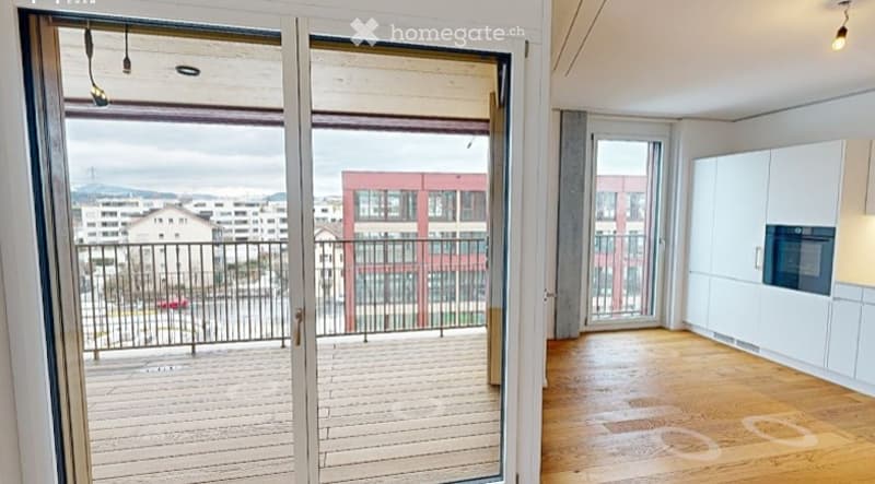 Modern, open-plan living space with a balcony overlooking a cityscape. The room features light wooden flooring, white kitchen cabinets, and a built-in oven. The balcony has a wooden deck and metal railing.