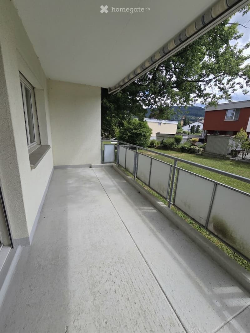 Balcony with concrete floor, white walls, metal railing, and a view of a green garden and surrounding buildings.