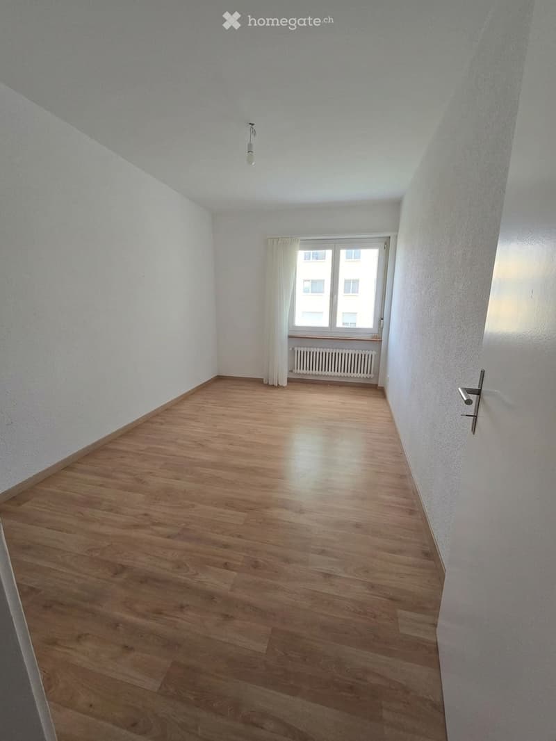 Empty room with wooden flooring, white walls, a window with white curtains, and a radiator below the window. No furniture or equipment is present.