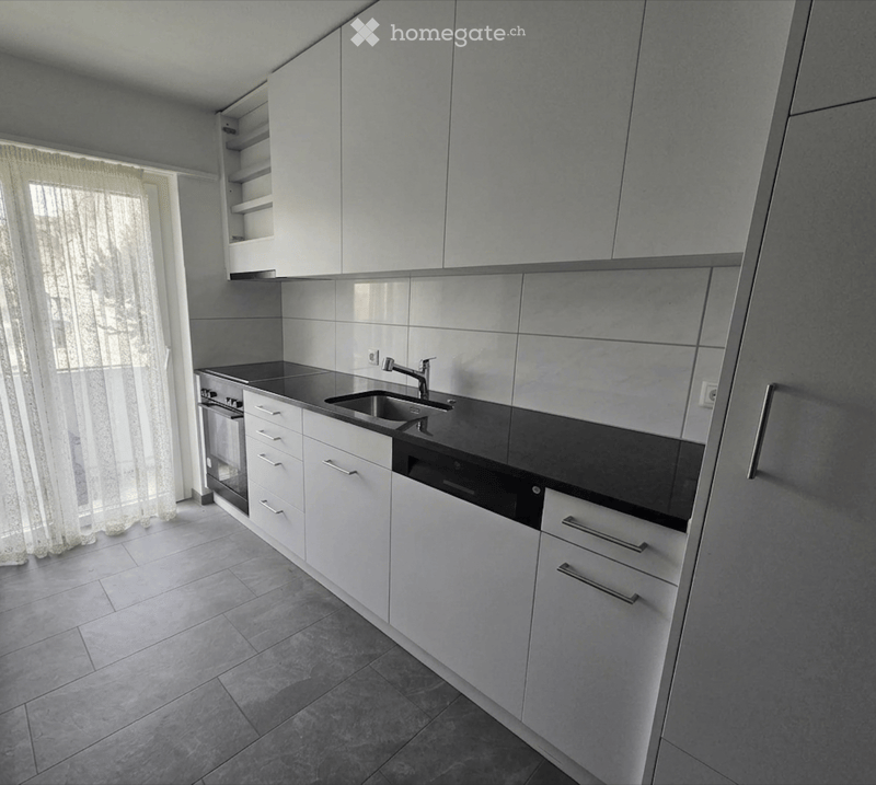 Modern kitchen with white cabinets, stainless steel sink, and built-in oven. Light grey tiles and a window with sheer curtains.