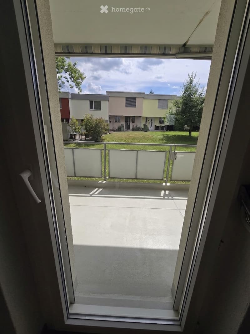 Balcony with a view of a residential area, featuring a metal railing and a sliding glass door with a striped blind.