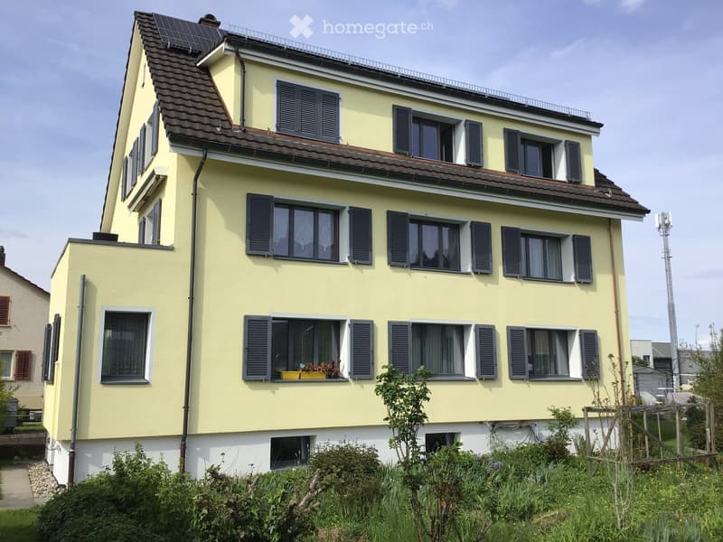 Two-story yellow house with multiple windows, shutters, and a small balcony on the roof. The house has a garden in front with various plants and shrubs.