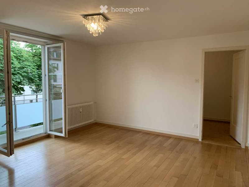 Empty room with wooden flooring, white walls, large sliding glass door leading to a balcony, and a radiator under the window.