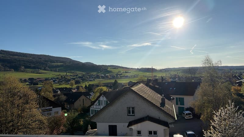 Residential neighborhood with houses, trees, and a scenic view of hills and fields under a clear blue sky.