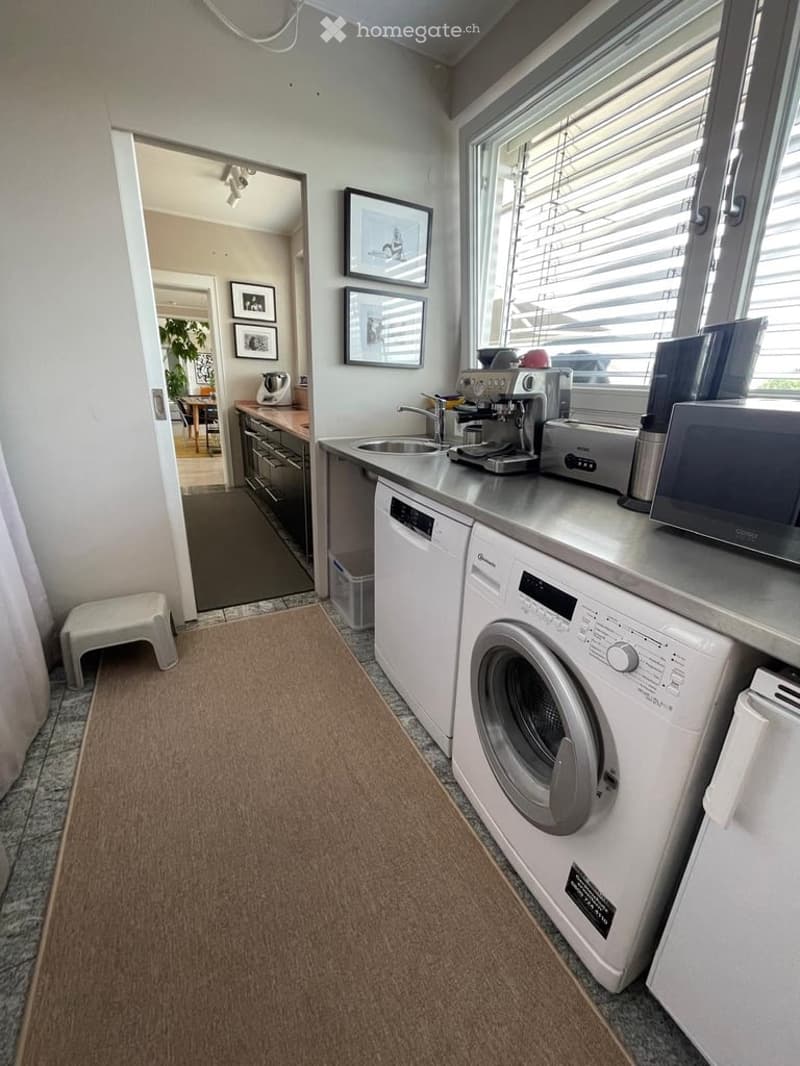 Modern kitchen with stainless steel appliances, including a sink, dishwasher, washing machine, coffee maker, and microwave. A small white stool is near the entrance, and framed pictures adorn the walls. The space is well-lit with natural light from a larg