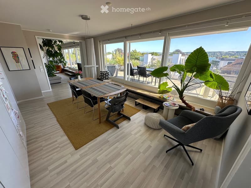Modern living room with wooden flooring, large windows offering a scenic view, and various seating arrangements including a sofa, chairs, and a small table.