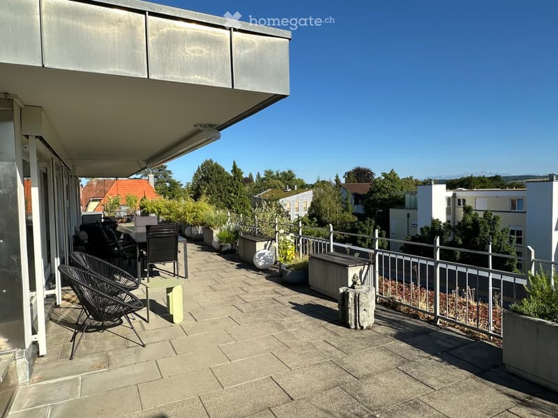 Balcony with tiled floor, metal railing, several chairs and a table, potted plants, and a view of surrounding buildings and greenery.