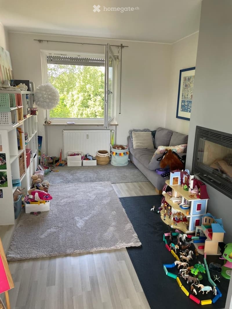Bright living room with a grey sofa, shelves filled with books and toys, a large window with blinds, and various children's toys scattered around, including a toy horse and a toy house.