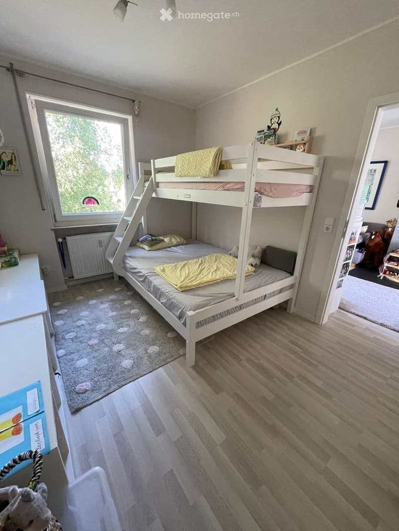Cozy bedroom with a wooden bunk bed, nightstand, and a small rug on the floor. The room features wooden flooring and a large window with a view of greenery.