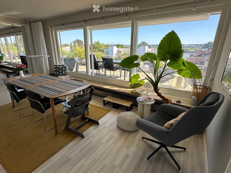 Modern living room with large windows, wooden floor, grey sofa, wooden table, chairs, small bench, round stool, and decorative plant.