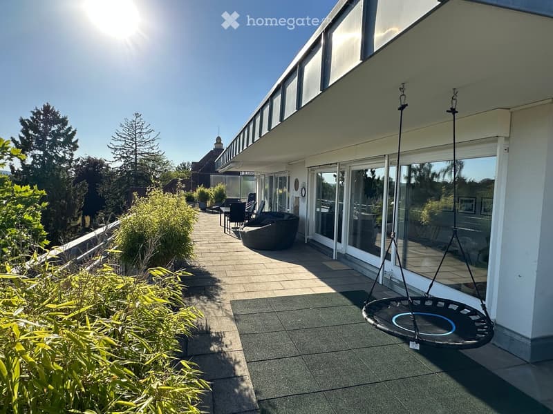 A modern outdoor patio area with a trampoline, seating area, and lush greenery, connected to a house with large glass windows and a tiled walkway.