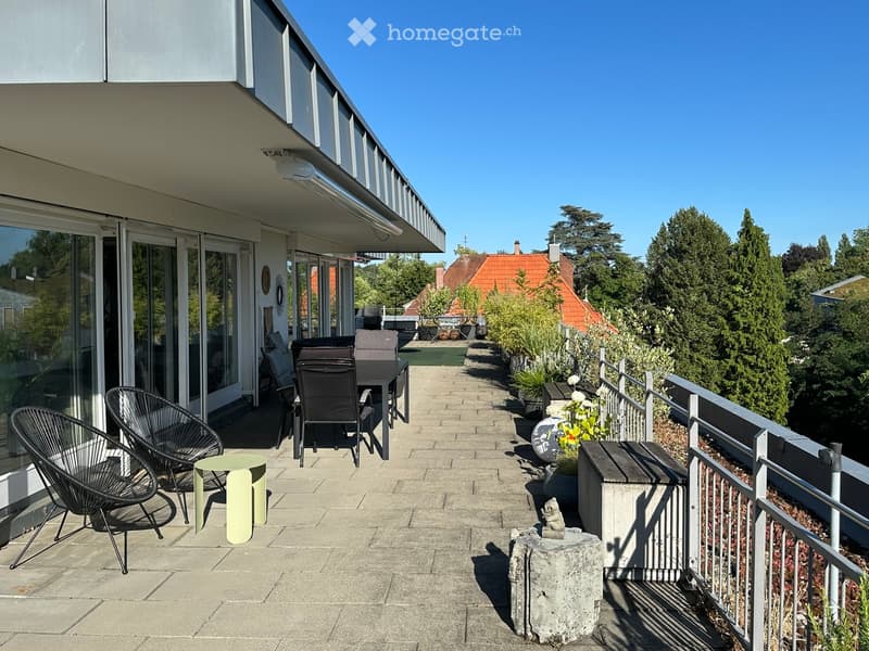 Balcony with modern black chairs, a small green table, and a bench, surrounded by greenery and a clear blue sky.