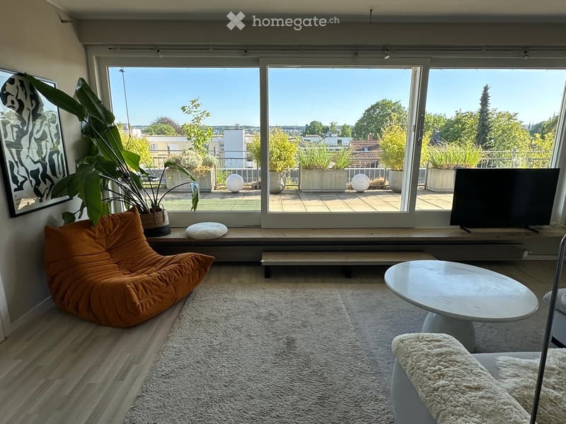 Modern living room with large windows offering a scenic view, featuring a round white coffee table, an orange bean bag sofa, a TV screen, and a potted plant near the window.
