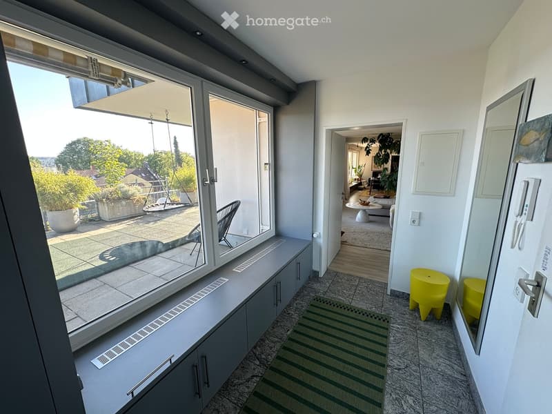 Bright hallway with large windows offering a view of the outdoor area, featuring a green and white striped rug, yellow stools, and a mirror on the wall.