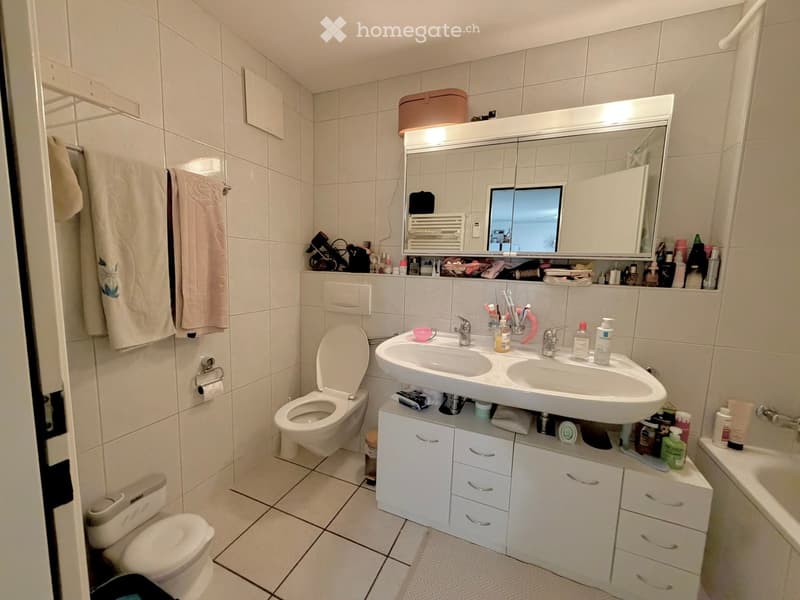 A modern bathroom with white tiles, a double sink, a toilet, a bathtub, and a large mirror with built-in storage. The sink area has a cabinet with multiple drawers and shelves holding various toiletries.
