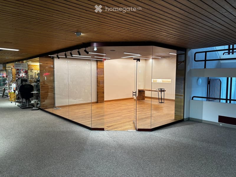 Modern hallway with wooden ceiling, glass partitions, and carpeted floor. Features a small table and chairs in one section, and a clothing display area visible through the glass.