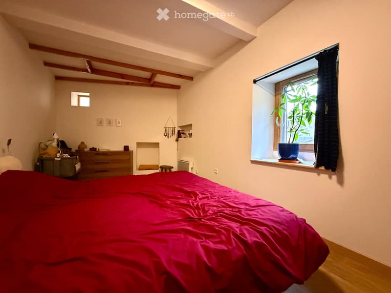 Cozy bedroom with a large bed covered in a red duvet, a wooden dresser, a small stool, a mirror, and a window with a potted plant on the sill.