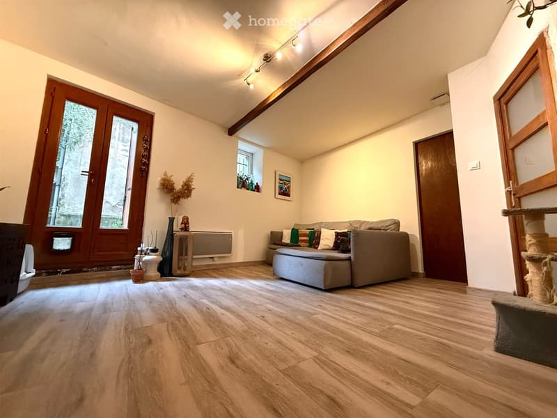 Cozy living room with wooden flooring, large window, grey sofa with colorful cushions, ottoman, small table with vase, and cat tree.