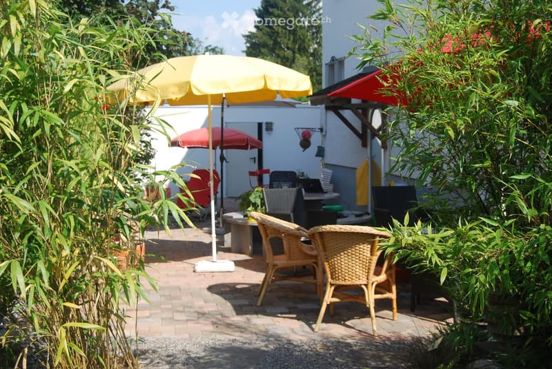 A sunny outdoor patio area with wicker chairs, umbrellas, and a small table surrounded by lush greenery and a modern house in the background.