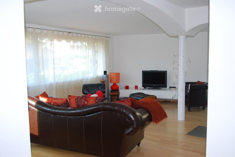 Modern living room with a brown leather sofa, red throw, armchairs, a small table with a red lamp, and a television on a white stand.