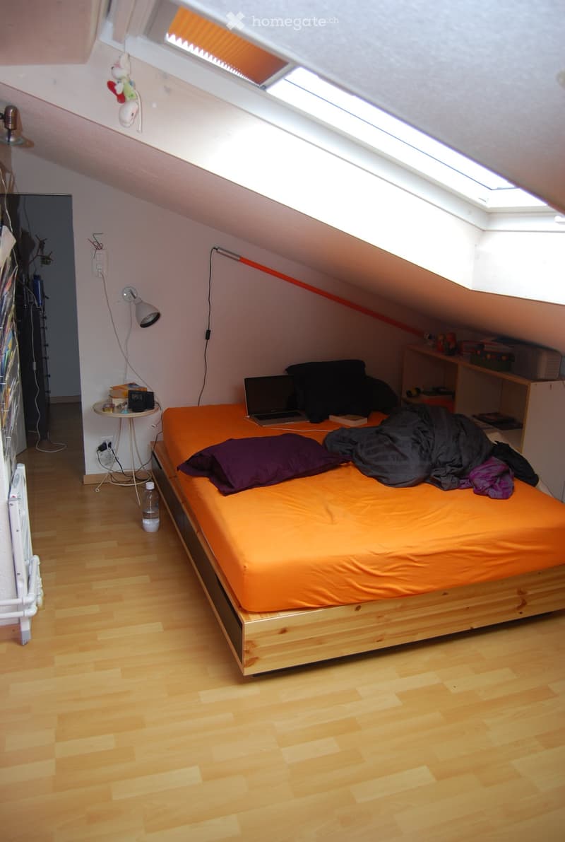Cozy attic bedroom with wooden floors, slanted ceilings, and skylights. Features a single bed with orange bedding, a small table with a lamp, and a shelf with various items.