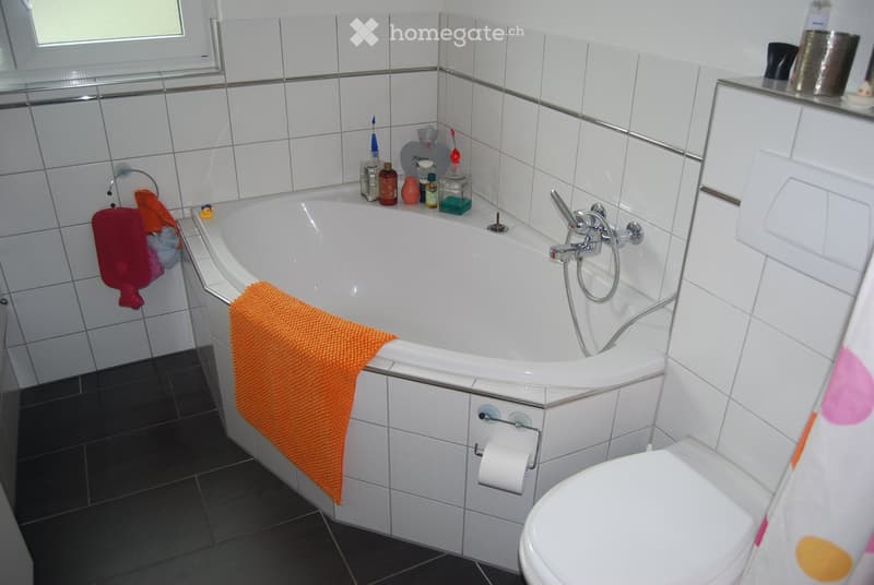 Bathroom with white tiled walls, a bathtub with an orange towel draped over the edge, a toilet, and various toiletries on the edge of the tub.