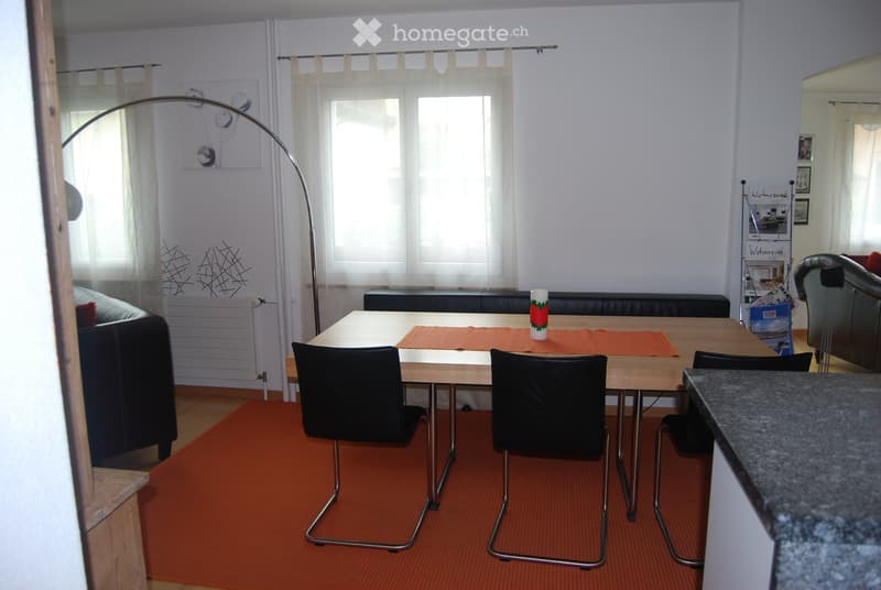 Bright living room with a wooden dining table, four black chairs, a sofa, a floor lamp, and a cup on the table. The room features large windows with white curtains and an orange carpet.