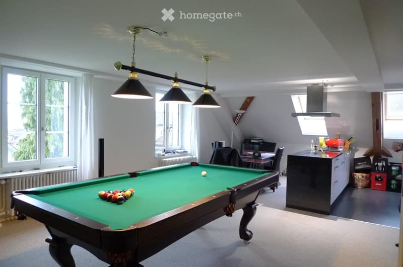 A spacious room with a pool table, surrounded by large windows and modern lighting. A kitchen area is visible in the background with cabinets and a counter.
