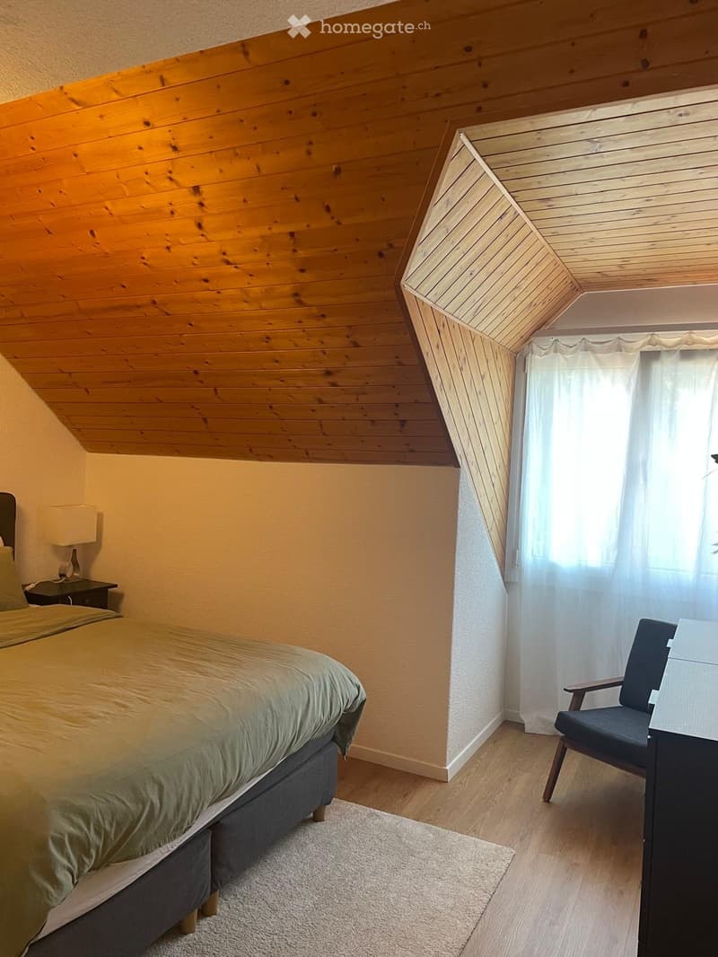 Cozy bedroom with wooden slanted ceiling, bed with green bedding, wooden nightstand with lamp, and a chair near the window with white curtains.
