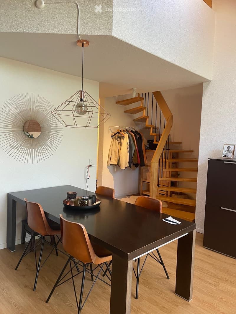 Dining area with a dark wooden table, four modern chairs, a hanging geometric lamp, a sun-shaped wall mirror, and a staircase leading to another level.