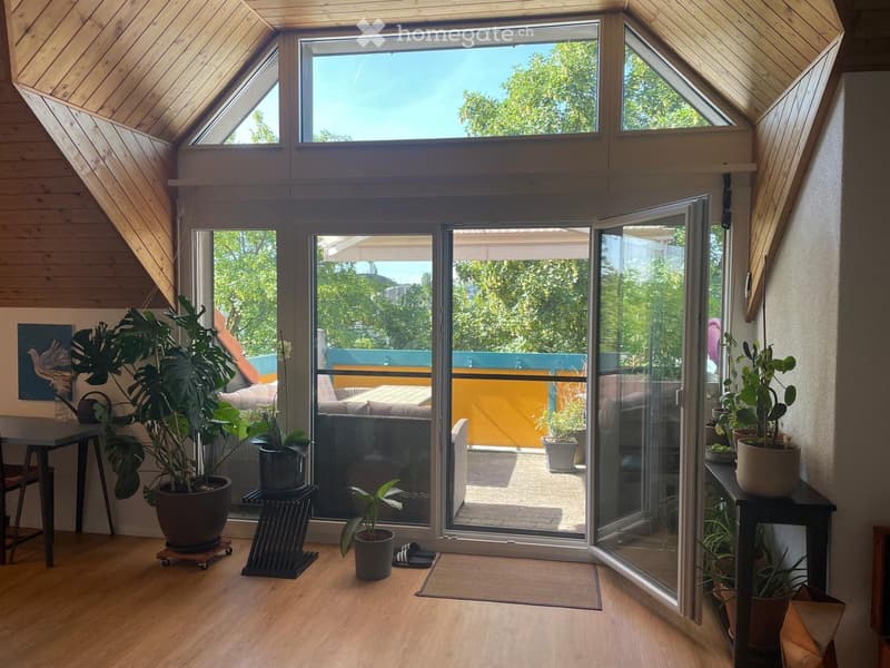 Bright, airy room with wooden floors, large windows, and sliding glass doors leading to a balcony. Features include a table, chairs, potted plants, and a sofa visible through the glass doors.
