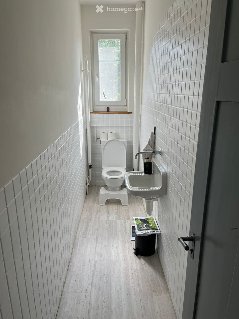 Bathroom. A narrow bathroom with white tiled walls, a toilet, a sink with a faucet, a soap dispenser, a small window, and a magazine on the floor next to a trash can.