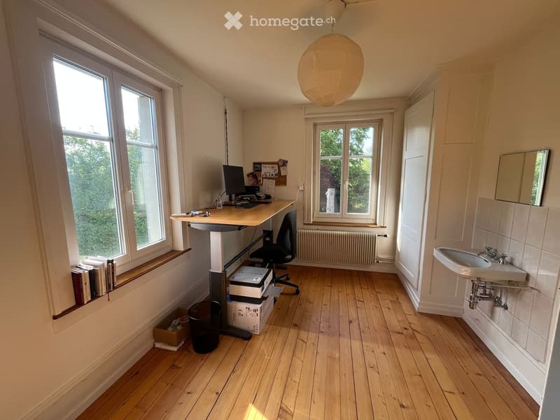 Furnished Room. A furnished room with a desk, chair, computer, sink, mirror, radiator, books, printer, and a lamp. The room has wooden flooring and large windows with a view of greenery.