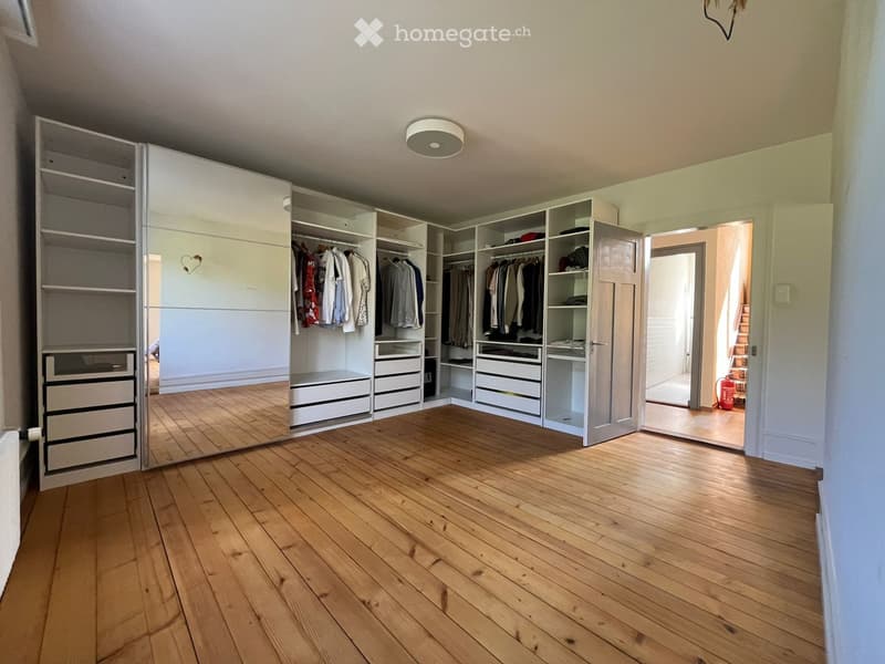 Unfurnished Room. Empty room with wooden flooring, large built-in wardrobe with multiple sections, drawers, and shelves. The wardrobe is partially open, revealing hanging clothes and storage compartments. A small heart-shaped decoration is visible on the