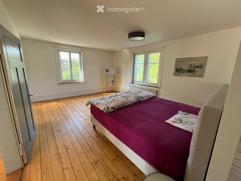 Bedroom. Cozy bedroom with wooden flooring, a double bed with a purple duvet and decorative pillows, a towel rack, a painting on the wall, and a radiator under the windows.