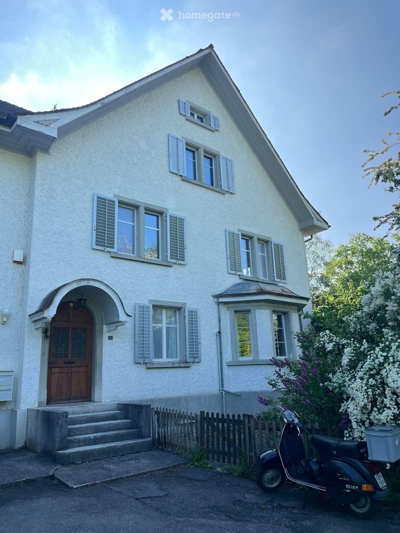 House. Two-story white house with multiple windows, shutters, and a wooden front door. A scooter is parked in front of a wooden fence with flowering bushes.