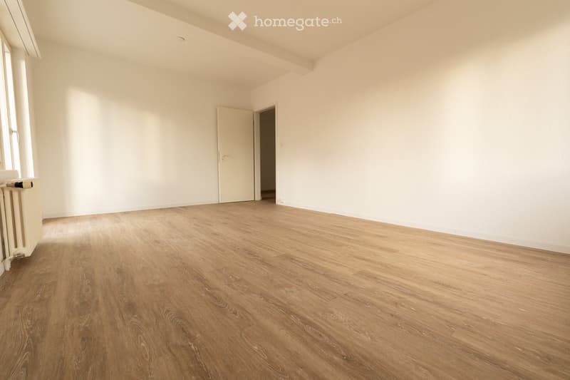 Empty room with light wooden flooring, white walls, and a single door leading to another space. Natural light streams in from a window on the left side.