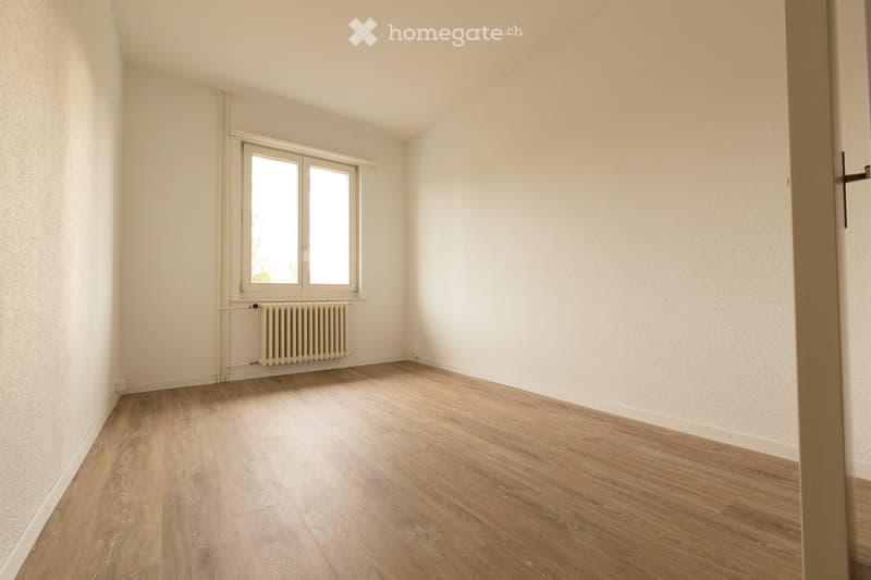 Empty room with light wooden flooring, white walls, and a window with a radiator below it.