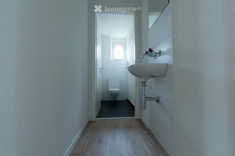 A narrow hallway with light wooden flooring and white walls, leading to a small bathroom with a toilet, sink, and mirror.