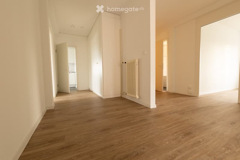 Empty hallway with wooden flooring, white walls, and a radiator on one side. Doors lead to adjacent rooms.