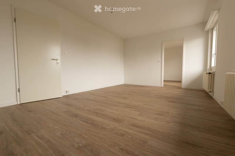 Empty room with wooden flooring, white walls, and a door leading to another space. Radiators are visible along the wall.