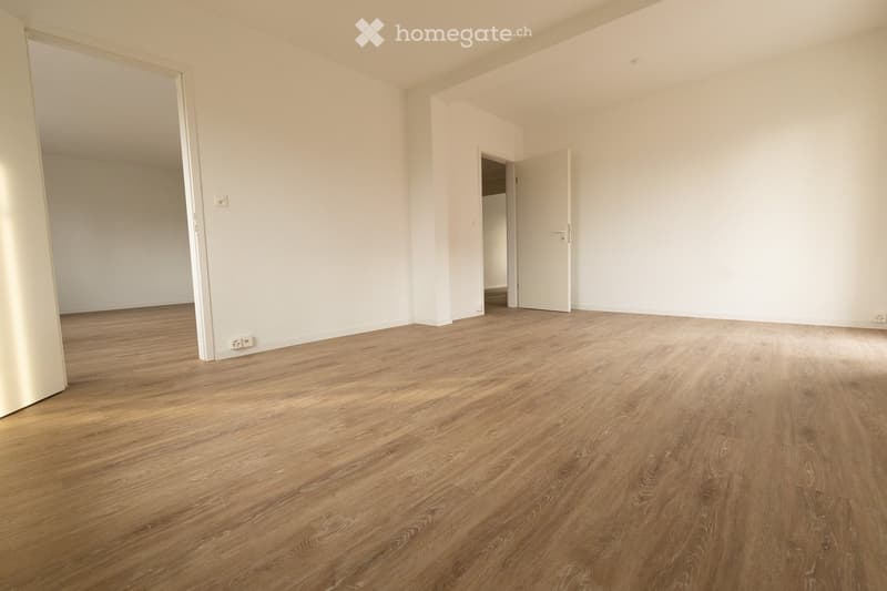Empty room with light wooden flooring and white walls, featuring two open doors leading to adjacent spaces.