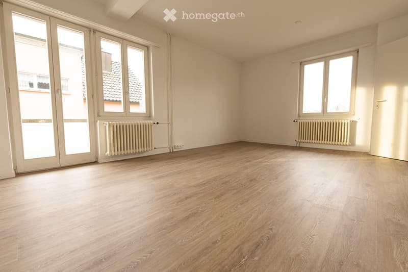 Empty room with light wooden flooring, white walls, large windows, and radiators on both sides.