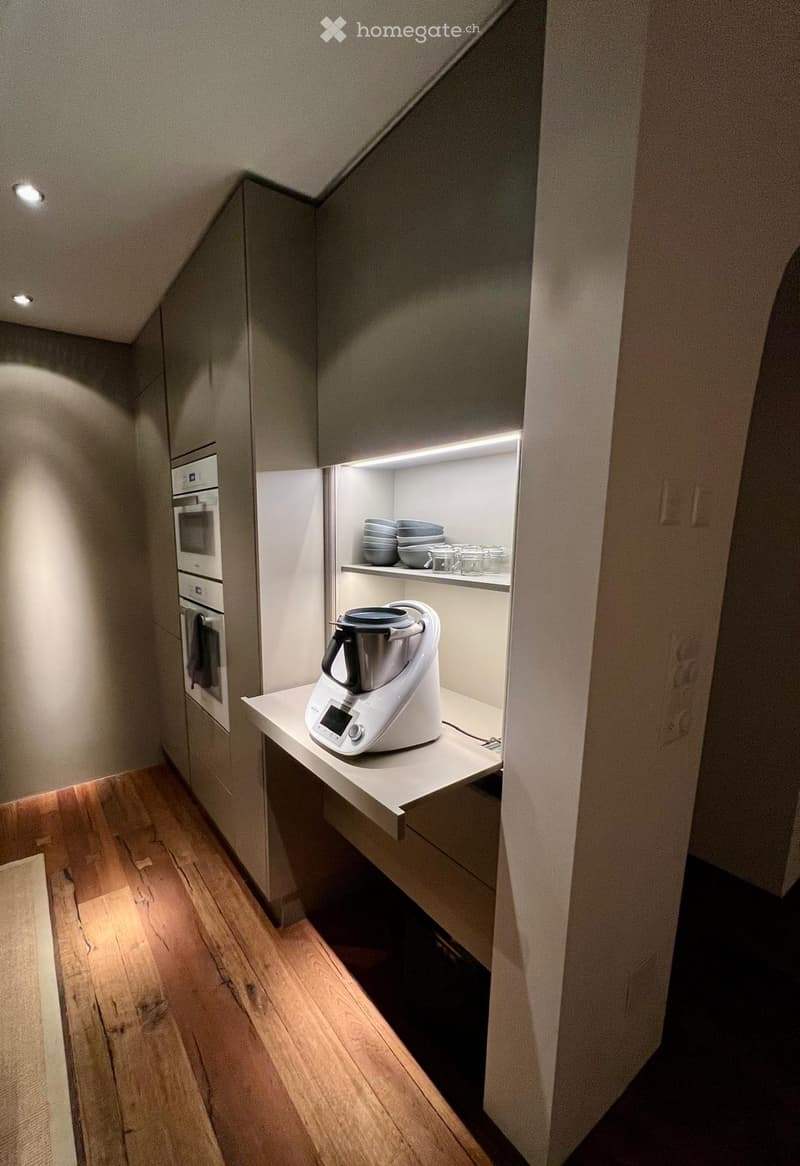 Modern kitchen with wooden flooring, built-in oven, countertop with a blender, and open shelving holding bowls and glasses.