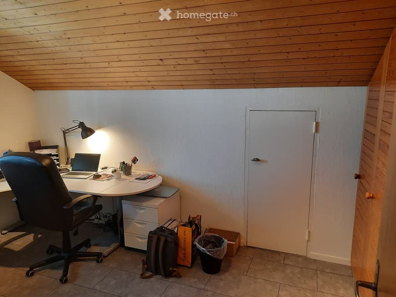 A small, well-lit room with a wooden ceiling and tiled floor. It features a desk with a laptop, a chair, a lamp, and various small items. A door is visible on the right side.