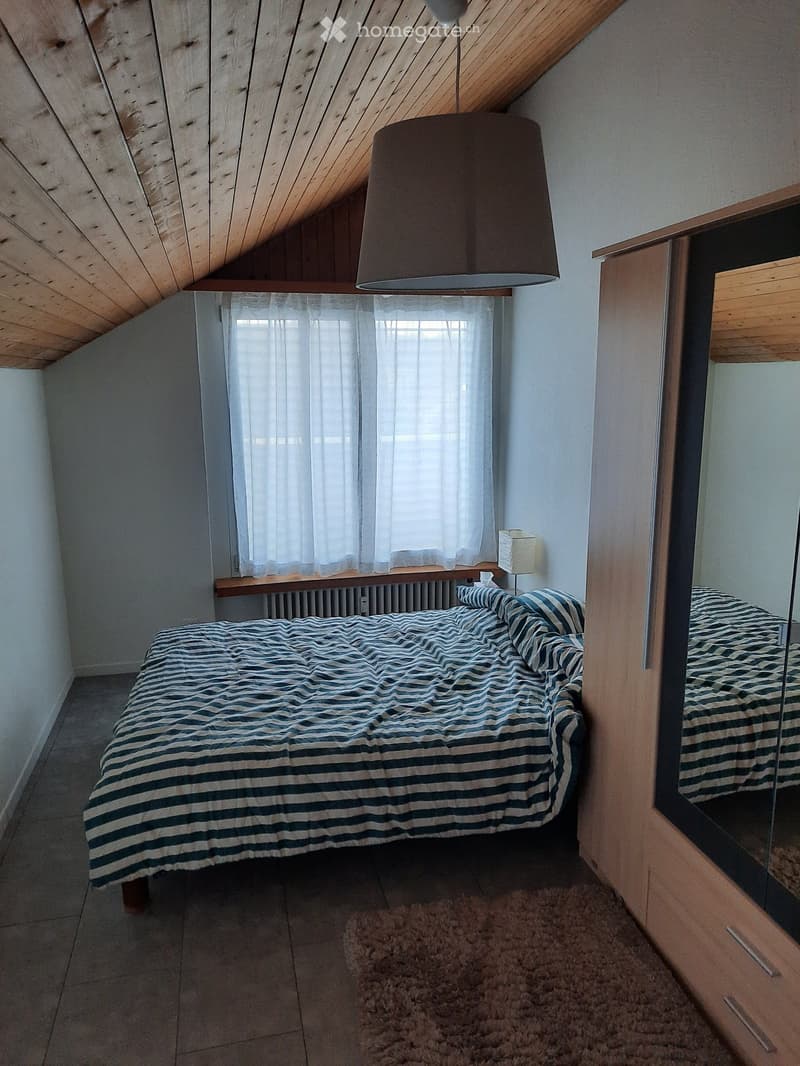 Cozy attic bedroom with slanted wooden ceiling, large window with white curtains, bed with blue and white striped bedding, mirror, and a small lamp on the nightstand.