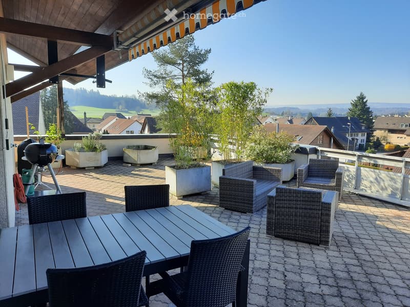 Balcony with a wooden table, wicker chairs, grill, and potted plants, overlooking a scenic countryside view.